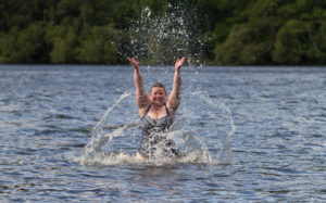 Photo of a woman in a loch in Scotland. She is wearing a swimming costume and is up to her waist in water. She is splashing water.