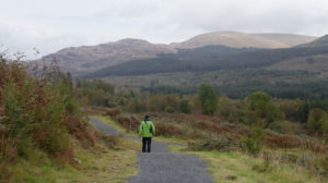 Image of a man walking down a woodland trail at Glen Trool. Hills in the background