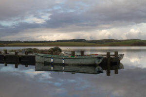 Image of a small boat moored on a jetty. The reflections of the clouds on the water