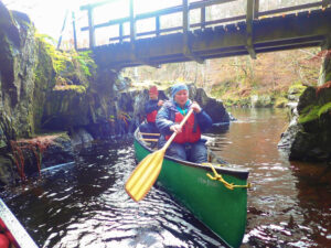Photo of and man and a woman canoeing under a small wooden footbridge